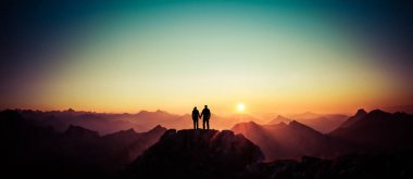 Happy winning success man and woman at sunset or sunrise standing on mountain peak holding hands and are happy for having reached mountain top summit goal during hiking travel trek. Tirol, Austria
