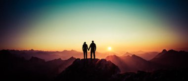 Happy winning success man and woman at sunset or sunrise standing on mountain peak holding hands and are happy for having reached mountain top summit goal during hiking travel trek. Tirol, Austria