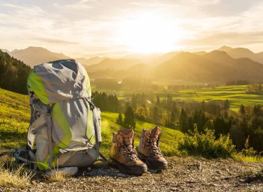 Hiking equipment in early morning sunrise and mountain silhouettes. Backpack and hike shoes in beautiful sunlight and sunbeams. Allgaeu, Bavaria, Alps, Germany.