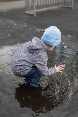 Yağmurdan sonra su birikintisinde oynayan yağmurlu pantolonlu ve lastik çizmeli iki yaşında bir çocuk.