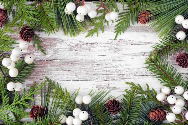 Winter frame with evergreen branches, white berries and pine cones. Top down view on a rustic white wood banner background with copy space.