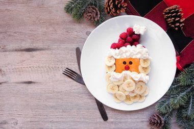 Cute Christmas Santa Claus pancake on a white plate with raspberry hat and banana beard. Top down table scene against a wood background. clipart