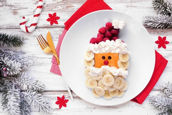 Fun Christmas Santa Claus pancake on a white plate with raspberry hat and banana beard. Overhead view table scene against a white wood background.