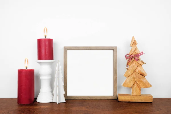 stock image Christmas mock up with wooden frame, tree decor and red candles. Square frame on a wood shelf against a white wall. Copy space.