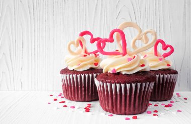 Valentines Day theme red velvet cupcakes with sweet hearts and white icing. Side view with a white wood background.