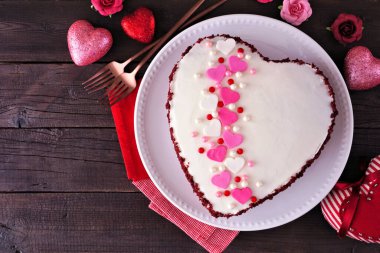 Valentines Day theme red velvet heart shaped cake with white icing and colorful heart decoration. Top view over a dark wood background.