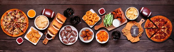 Superbowl or football theme food table scene. Pizza, hamburgers, wings, snacks and sides. Above view on a dark wood banner background.