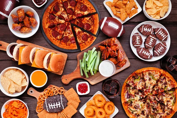 stock image Super Bowl or football theme food table scene. Pizza, hamburgers, wings, snacks and sides. Overhead view on a dark wood background.