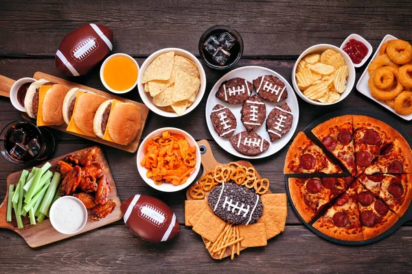 Super Bowl ya da futbol temalı yemek masası sahnesi. Pizza, hamburger, kanat, aperatif ve meze. Koyu ahşap arka planda üst görünüm.