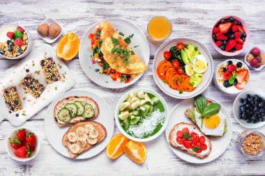 Healthy breakfast food table scene. Above view over a white wood background. Omelette, nutritious bowl, toasts, granola bars, smoothie bowl, yogurts and fruits.