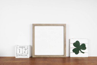 Mock up wood frame with St Patricks Day decor on a wood shelf. Shabby chic wood sign and calendar. Square frame against a white wall. Copy space. clipart