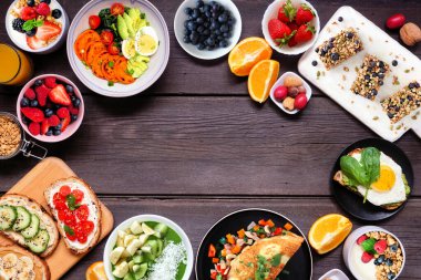 Healthy breakfast food frame. Top view over a dark wood banner background. Table scene with omelette, nutritious bowl, toasts, granola bars, smoothie bowl, yogurts, fruit. Copy space.