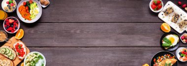 Healthy breakfast food double border. Top down view over a dark wood banner background. Table scene with omelette, nutritious bowl, toasts, granola bars, smoothie bowl, yogurts, fruit. Copy space.