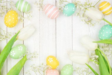 Pastel Easter eggs and white spring tulip flowers. Overhead view frame against a white wood background. Copy space.