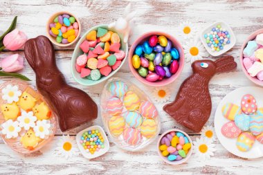 Easter candy table scene. Top down view over a white wood background. Chocolate bunnies, candy eggs and a collection of sweets.