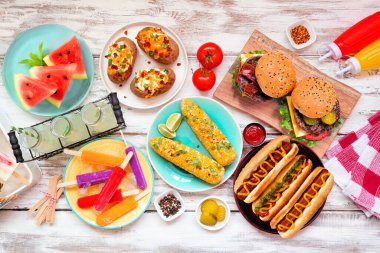 Yaz BBQ yemek masası sahnesi. Hamburger, sosisli sandviç, patates, mısır ve soğuk ikramlar. Beyaz ahşap arka plan üzerinden genel görünüm.