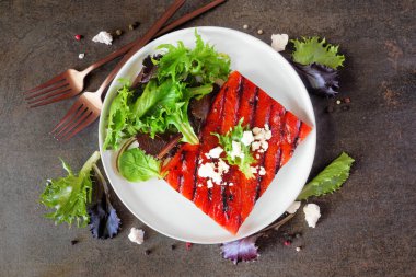 Izgara karpuz bifteği ve salata. Koyu taştan bir arka planda malzeme çerçevesi olan yukarıdan aşağıya doğru bir manzara. Sağlıklı beslenme, bitki temelli et ikame konsepti.