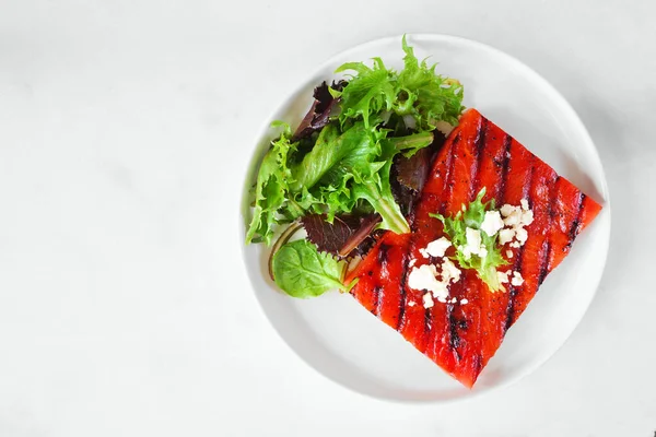 stock image Grilled watermelon steak with salad. Top view on a white marble background. Healthy eating, plant based meat substitute concept.