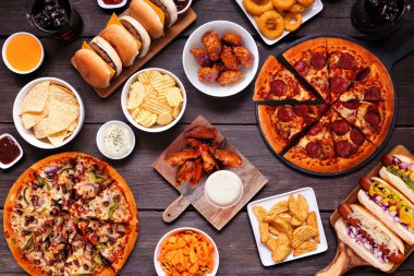Abur cubur masası sahnesi. Pizza, hamburger, sosisli sandviç, tavuk kanadı ve tuzlu atıştırmalıklar. Koyu bir ahşap arka plan üzerinden genel manzara.