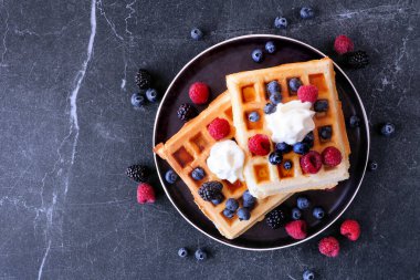 Ahududu, yaban mersini ve kremalı kahvaltı waffle 'ları. Koyu renkli bir arkaplan üzerinde üst görünüm.