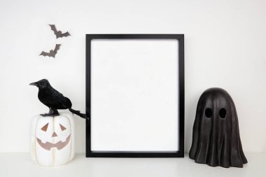 Cadılar Bayramı modeli. Beyaz bir rafta siyah çerçeve içinde Jack O fener, hayalet ve karga dekoru var. Beyaz bir duvara dayalı portre çerçevesi. Boşluğu kopyala.