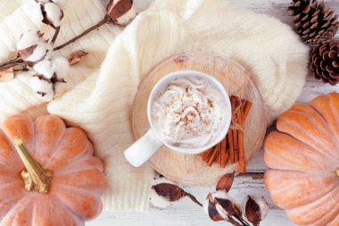 Balkabakları, süveter, doğal dekor ve balkabağı aromalı latte ile sıcacık sonbahar masası sahnesi. Beyaz ahşap arka planda tepeden aşağı görünüm.