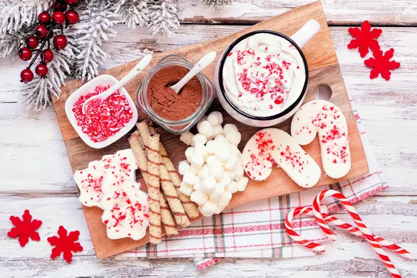 Christmas hot chocolate tray a variety of sweet toppings. Above view table scene on a white wood background.
