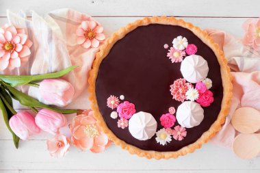 Kremalı çikolatalı tart ve pembe çikolata çiçekleri. Beyaz ahşap bir arka planda, yukarıdaki masa manzarası. Bahar pişirme konsepti.