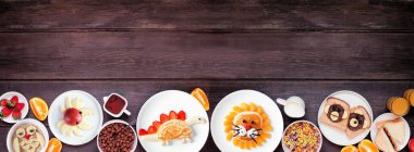 Eğlenceli çocuk temalı kahvaltı temalı alt sınırda çeşitli hayvan temalı yiyecekler. Koyu ahşap arka planda üst manzara. Gözleme, yulaf ezmesi, tost, meyve ve tahıl.