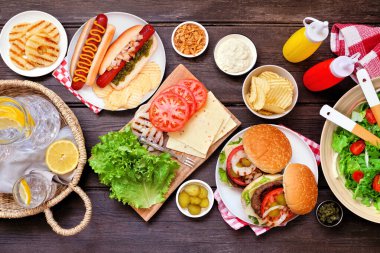 Yaz barbeküsü ya da hamburger, sosisli sandviç, salata ve abur cuburlu piknik masası sahnesi. Koyu bir ahşap arka planda manzaranın üstünde.