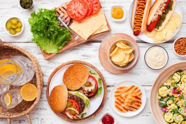 Yaz barbeküsü ya da hamburger, sosisli sandviç, salata ve abur cuburlu piknik masası sahnesi. Beyaz ahşap arka plan üzerinden genel görünüm.