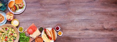 Yaz barbeküsü ya da hamburger, sosisli sandviç, salata ve abur cuburla piknik köşesi sınırı. Koyu ahşap bir arkaplanda manzaranın üstünde. Boşluğu kopyala.