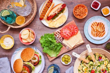 Yaz barbeküsü ya da hamburger, sosisli sandviç, salata ve abur cuburlu piknik masası sahnesi. Koyu bir ahşap arkaplan üzerinden yukarıdan aşağı görünüm.