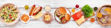 Yaz barbeküsü ya da hamburger, sosisli sandviç, salata ve abur cuburlu piknik masası sahnesi. Beyaz ahşap bir arkaplan üzerinde üst görünüm.