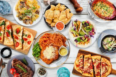 Çeşitli lezzetli yiyeceklerin olduğu masa sahnesi. Beyaz ahşap arka planda manzaranın üstünde. Makarna, ekmek, taco, biftek ve restoran temalı yemekler..