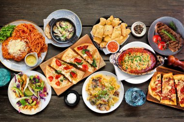 Çeşitli lezzetli yiyeceklerin olduğu masa sahnesi. Koyu bir ahşap arka planda tepeden aşağı manzara. Makarna, ekmek, taco, biftek ve restoran temalı yemekler..
