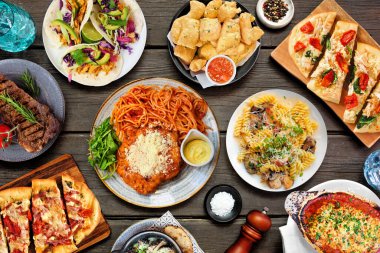 Çeşitli lezzetli yiyeceklerin olduğu masa sahnesi. Koyu bir ahşap arka planda tepeden aşağı manzara. Makarna, ekmek, taco, biftek ve restoran temalı yemekler..