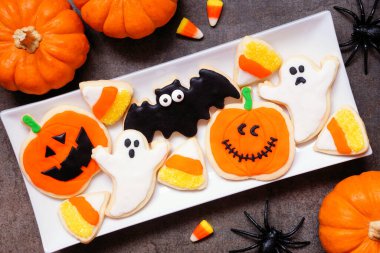 Bir tabak dolusu ürkünç Cadılar Bayramı kurabiyesi. Koyu masa arkaplanında balkabağı dekoru olan üst görünüm.