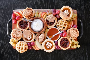 Sonbahar turtası temalı tatlı kurulu. Masanın üstünde, siyah ahşap bir arka planda. Balkabağı, elma ve cevizli mini turtalar, krem şanti, karamel ve meyveli..