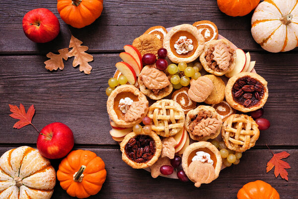 Осенний тематический десерт. Сцена стола сверху на фоне темного дерева. Тыквенные, яблочные и ореховые мини-пироги, печенье, тыквенные роллы и фрукты.