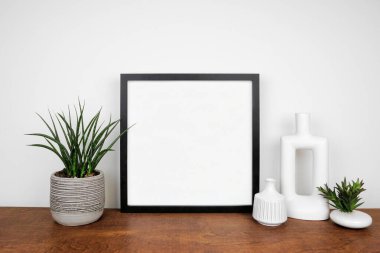 Siyah kare çerçeve, saksı bitkileri ve modern ev dekoruyla süslenir. Beyaz bir duvara karşı ahşap raf. Boşluğu kopyala.