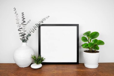 Siyah kare çerçeve, saksı bitkileri ve modern ev dekorlarıyla süslenir. Beyaz bir duvara karşı ahşap raf. Boşluğu kopyala.
