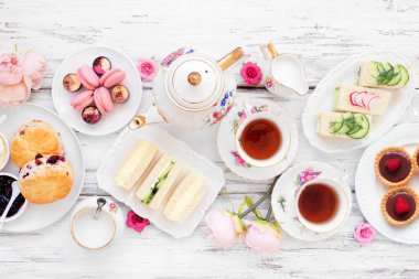 Akşamüstü çay masası sahnesi beyaz ahşap arka planda. Klasik çay seti, parmak sandviçler, çörekler ve tatlılar. Görünümün üstünde.