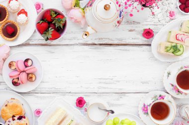 Akşamüstü çay çerçevesi beyaz bir ahşap zemine karşı. Klasik çay seti, parmak sandviçler, çörekler ve tatlılar. Kopyalama alanı olan üst görünüm.