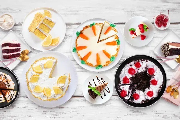 Pasta çeşitleriyle tatlı masası sahnesi. Beyaz ahşap arka planda üst manzara. Kara orman, limon, havuç, kırmızı kadife, çikolata ve peynirli kek..