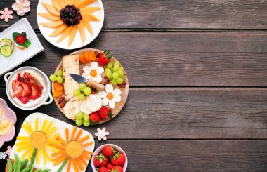 Yaz bahçesi partisi yiyecek sınırında. Çeşitli çiçek temalı şarküteri ürünleri, peynir, meyve, sebze ve parmak atıştırmalıkları. Koyu ahşap arka planda üst görünüm.