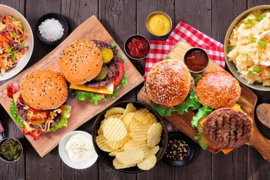 Hamburgerli barbekü masası sahnesi, klasik salatalar ve atıştırmalıklar. Koyu bir ahşap arka planda manzaranın üstünde.