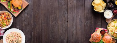 Hamburgerler, klasik salatalar ve abur cuburlarla barbekü. Masa manzarasının üstünde koyu ahşap bir afiş var. Boşluğu kopyala.