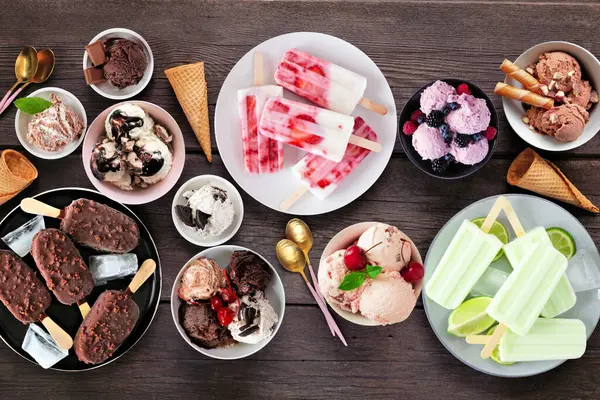 Harika bir yaz yemeği masası sahnesi. Çeşitli dondurma kaseleri, buzlu dondurmalar ve dondurulmuş ikramlar. Koyu ahşap arka planda üst görünüm.