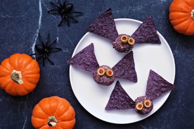 Cadılar Bayramı temalı aperatifler. Mor nacholu yarasa peyniri topları. Siyah taştan bir arkaplan karşısında yukarıdaki görünüm tablosu.